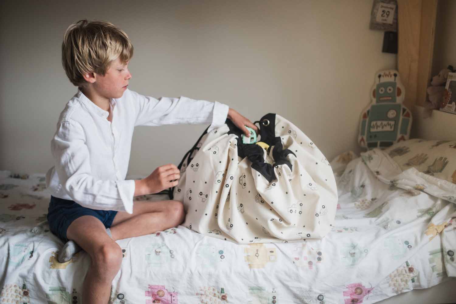 toy storage bag, cars print, child sitting on bed, putting toys in the bag
