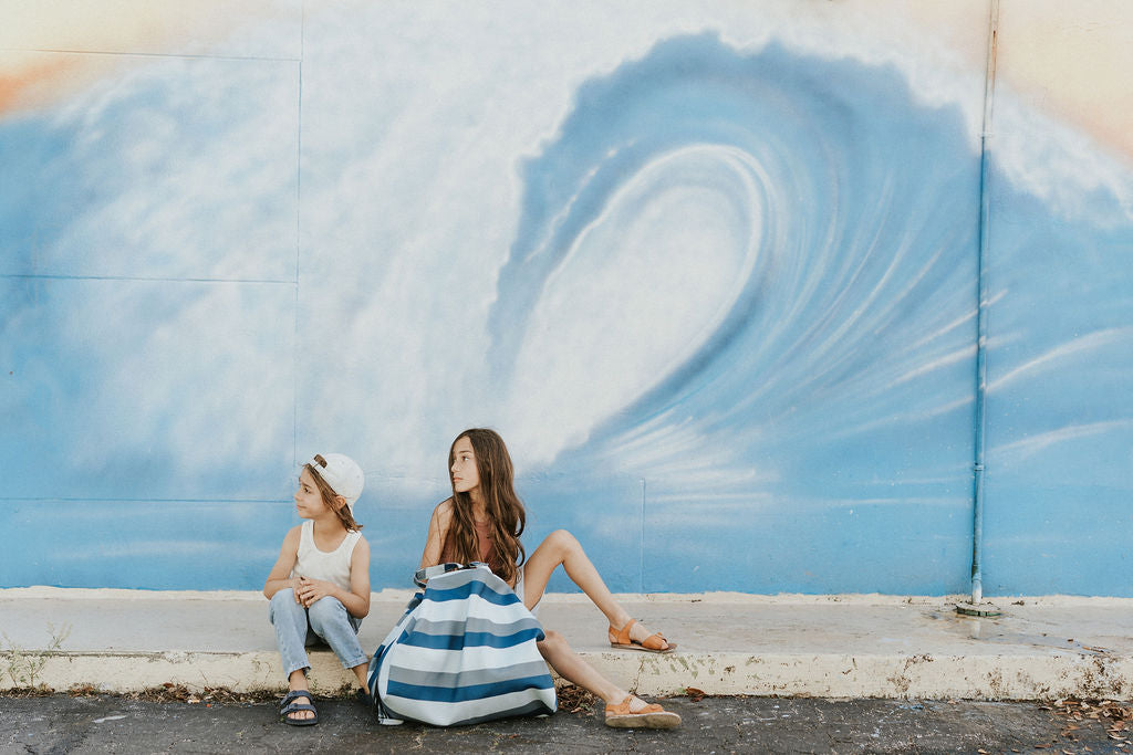 outdoor beach bag blue/green stripes, children sitting on the sidewalk with bag, big painted wave at the back on the wall