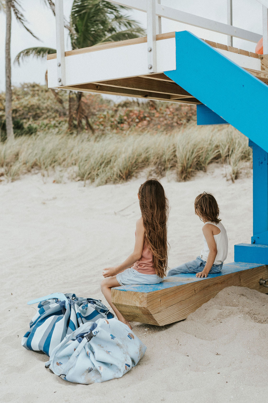 outdoor beach bag blue/green stripes and little boat print, 2 bags closed, next to each other, on the beach, children looking at the  grass                             