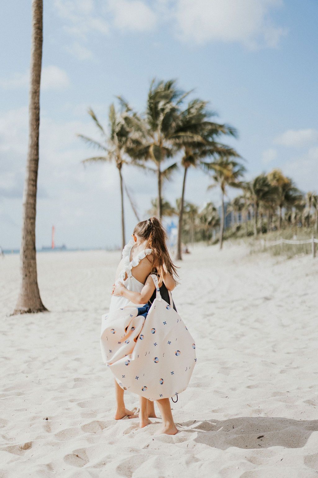 outdoor balloon storage bag and play mat, child carrying the bag on it's shoulder, lots of palm trees at the beach, children hugging each other