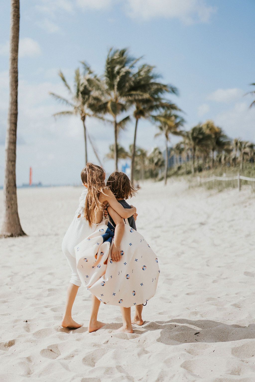 outdoor balloon storage bag and play mat, child carrying the bag on it's shoulder, lots of palm trees at the beach, children hugging each other