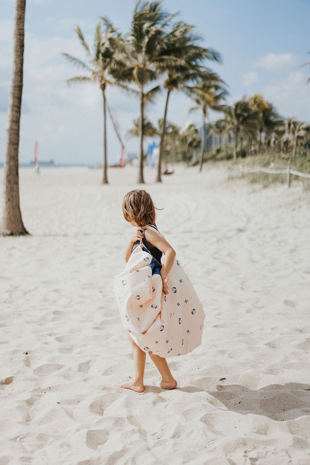 outdoor balloon storage bag and play mat, child carrying the bag on it's shoulder, lots of palm trees at the beach