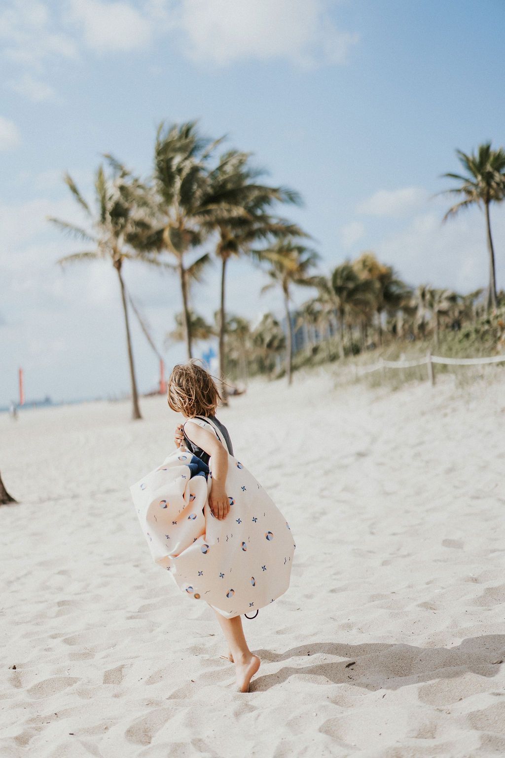 outdoor balloon storage bag and play mat, child carrying the bag on it's shoulder, lots of palm trees at the beach