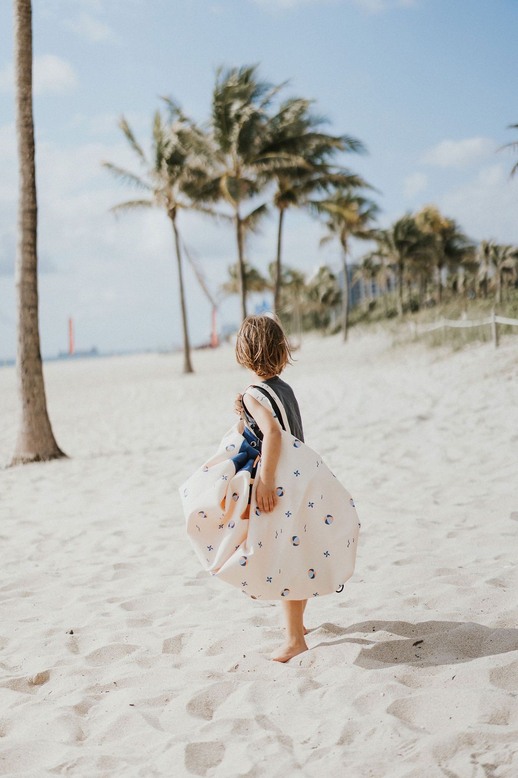 outdoor balloon storage bag and play mat, child carrying the bag on it's shoulder, lots of palm trees at the beach