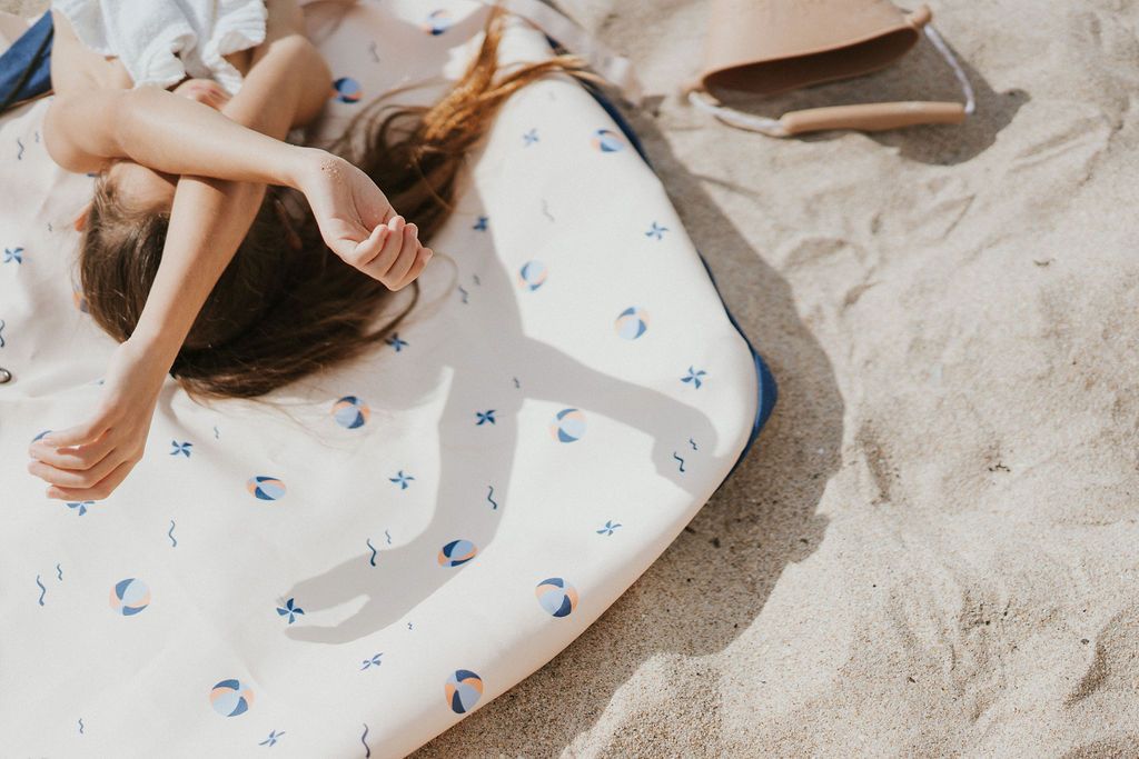 Play&Go outdoor storage bag and play mat, print balloon, child laying on open mat in the sand, holding arms before eyes